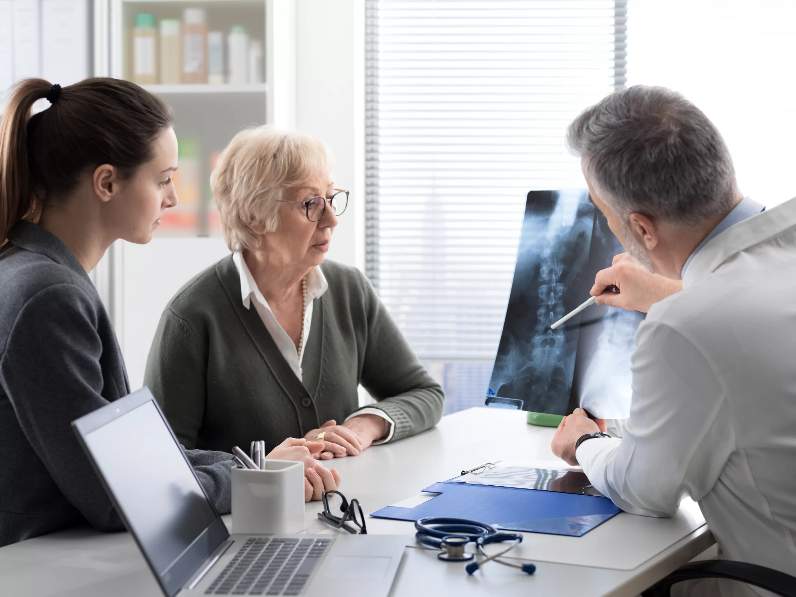 Physician Reviewing Spine X-Ray with Patient and Family Member Doctor explaining spinal X-ray results to elderly patient and her caregiver in a medical office.